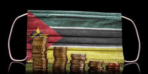 Medical mask with the flag of Mozambique behind some shrinking stacks of various coins.(series)
