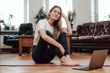 Beautiful woman sits on floor with laptop and smiles at home with modern design and styled interior.