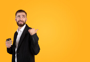 Positive young businessman in formal suit holding takeaway coffee and pointing at blank space on orange background