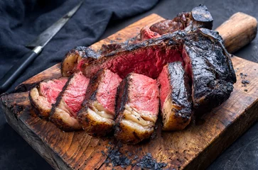 Tuinposter Traditional barbecue dry aged wagyu t-bone beef steak bistecca alla Fiorentina sliced and served with black salt as close-up on an old rustic wooden board © HLPhoto
