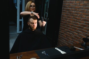 Obraz na płótnie Canvas Serving client in Barbershop. Professional barber girl, female hairdresser making modern haircut for a man sitting in barber shop chair. Focus on a girl. Hairdressing, shaving, trimming, grooming