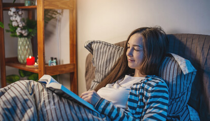 A young woman is reading a book in bed before going to bed in a dark bedroom. Concept of the...