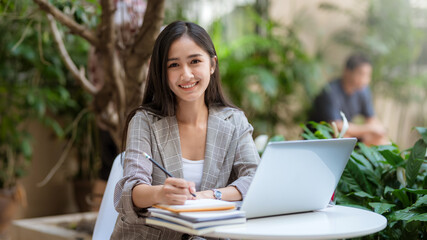 Asian businesswoman smiles at the camera while working in a casual laptop garden at a coffee shop.