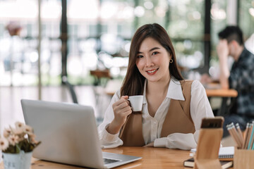 Fototapeta na wymiar Young Beautiful smiling Asian business woman holding a coffee and laptop Placed at the wooden table at the office