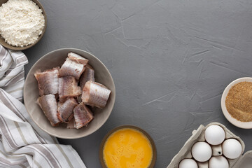 Portioned pieces of hake in a bowl and products for deep-fried cooking, eggs, flour and bread crumbs with a kitchen towel on a gray background