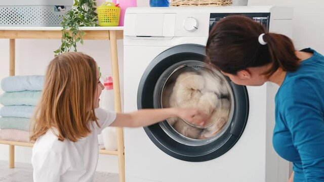 Mother with little daughter watching toys rolling in washing machine during laundry