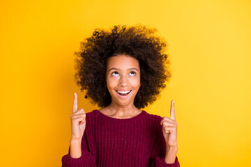 Portrait of astonished curly dark skin girl look curious direct fingers up empty space isolated on yellow color background
