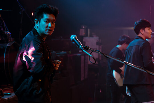 Singer Holding A Microphone Stand And Performing On Stage Looking At The Camera. Rock Concert Show Set Up.