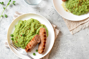 Pea mash and grilled sausages in a white ceramic plate on a gray concrete table top view. Tasty balanced lunch or dinner.