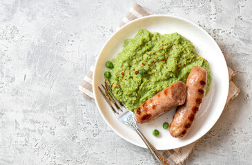 Pea mash and grilled sausages in a white ceramic plate on a gray concrete table top view. Tasty balanced lunch or dinner.