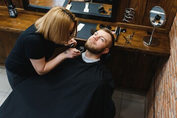 Skilled female barber cutting a beard of brunette adult guy in professional retro barbershop.