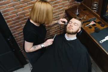Skilled female barber cutting a beard of brunette adult guy in professional retro barbershop.
