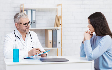 Concentrated senior man in white coat and glasses makes notes, looks at sad woman patient