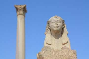 A sphinx in the northern Egyptian city of Alexandria with a column in the background. An ancient figure in the form of an animal with a human head. It is a clear day with blue skies.