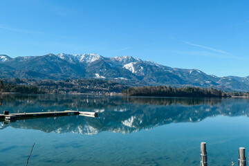 A panoramic view on the Faaker lake in Austrian Alps. The lake is surrounded by high mountains. Calm surface of the lake reflects the surrounding. A wooden pier going into the water. Clear, blue sky