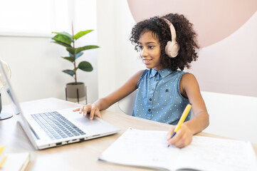 A pretty curly African schoolgirl wearing a headphones using a laptop for studying on the distance, a pretty biracial girl fill exercises book in with a pen, doing home work. Homeschooling concept
