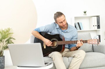 A young man is learning playing guitar himself, watching webinar, online masterclass, video tutorial. A guy holds acoustic guitar and repeat exercises, laptop in front of him. E-learning concept