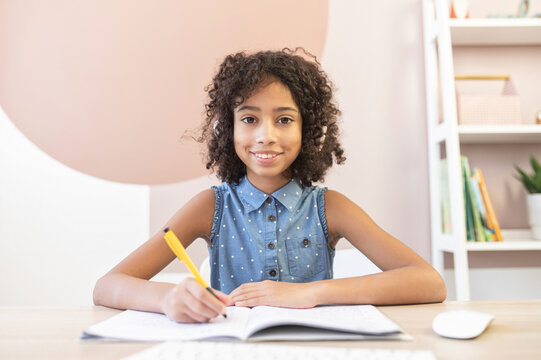 Webcam view of a pretty curly African schoolgirl wearing a headphones, a pretty biracial girl fill exercises book in with a pen watching online video classes. E-learning concept