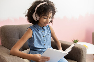 Clever and inquisitive African preteen school girl spending leisure time with a book reading sitting at armchair at home, preparing to exam