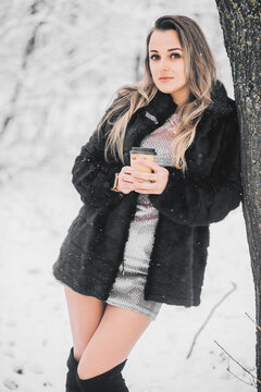 Beautiful young girl in a fur coat dress and high boots in the winter in the forest with coffee in her hands