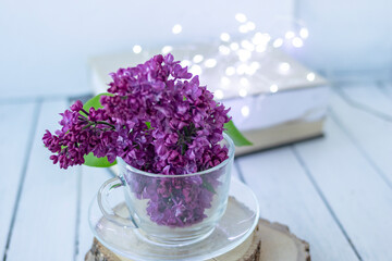 Transparent cup with flowers. Beautiful lilac fills the glass. The spring bouquet is worth no wooden cuts. Macro image of spring lilac purple flowers. Book in the background and lights. Blurred