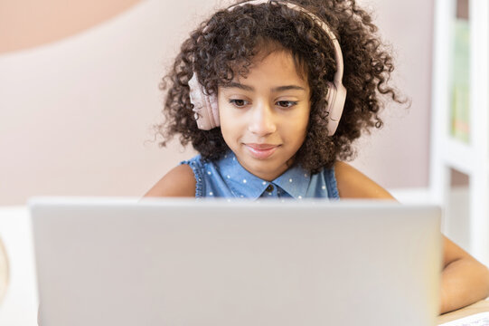 A pretty curly African schoolgirl wearing a headphones using a laptop for studying on the distance, a pretty biracial girl looks at the screen, doing homework. Homeschooling concept