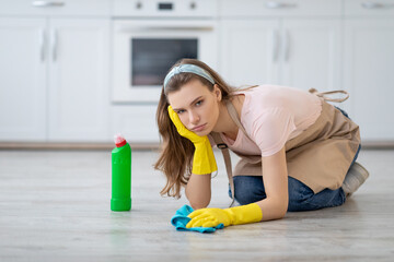 Upset young lady in rubber gloves and apron washing floor at kitchen, disliking house chores, feeling tired and unhappy