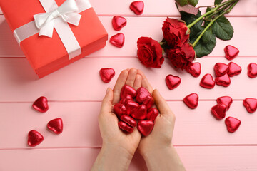 Woman holding heart shaped chocolate candies at pink wooden table, top view