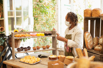 Female baker in face mask arranging pastry display in shop