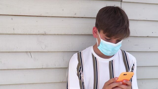 Teenage boy or young man outside wearing a face mask and using cell phone for social media during COVID-19 Coronavirus pandemic