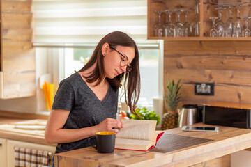 Woman relaxing at home reading a book and drinking coffee at kitchen counter