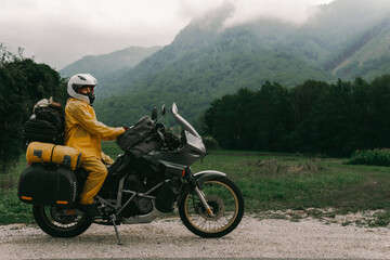 Fototapeta na wymiar A girl in a yellow raincoat, shoe covers and a white helmet. Motorcyclism and travel. Sightseeing tour. Top of the Mountains. A gray day with thunderclouds. Copy space, biker's outfit.