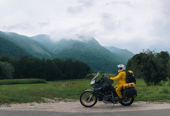 Fototapeta na wymiar A girl in a yellow raincoat, shoe covers and helmet. Motorcyclism and travel. Sightseeing tour. Top of the Mountains. A gray day with clouds. Copy space, biker's outfit. High resolution panorama