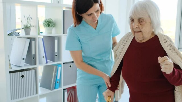 caregiver helping elderly woman in light room inside nursing home