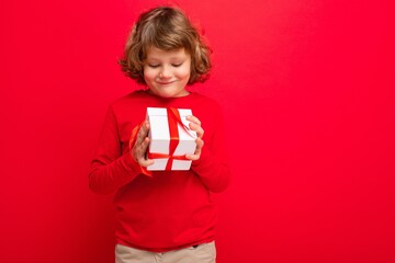 Positive blonde curly boy isolated over red background wall wearing red sweater holding gift box and looking at surprise box