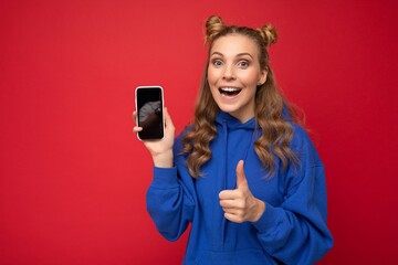 Photo of attractive crazy amazed surprised young woman wearing casual stylish clothes standing isolated over background with copy space holding and using mobile phone looking at camera giving thumbs