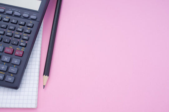 Overhead partial view of a scientific calculator, grid paper and pencil on a pink surface