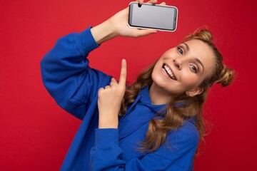 Naklejka premium Attractive smiling young blonde woman wearing stylish blue hoodie isolated on red background with copy space holding smartphone showing phone in hand with empty screen display pointing at gadjet