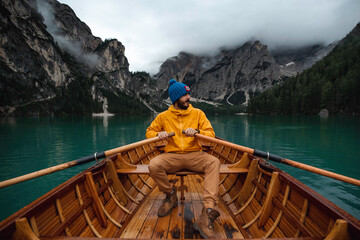 Traveler man with blue hat and yellow raincoat sailing in a wooden boat through a beautiful Di Braies lake in the Dolomites mountains. 