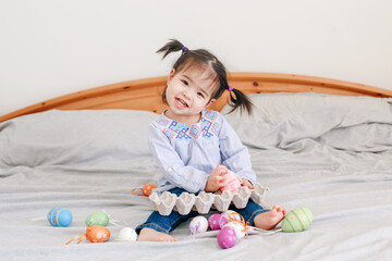 Obraz premium Happy Easter. Asian Chinese baby girl with funny pigtails sitting on bed at home and playing with colorful Easter eggs. Kid child celebrating a traditional Christian holiday.
