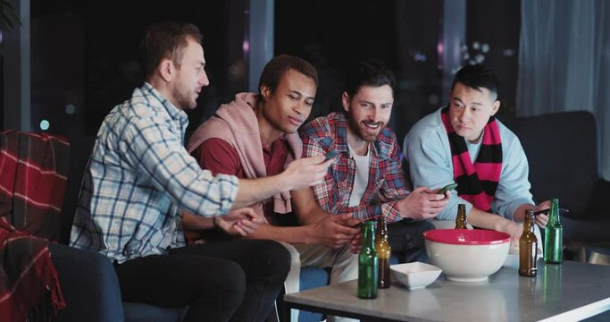 Multi-race young people using smartphones watching TV at night together. Diverse men football fans cheering for team sitting in living room.