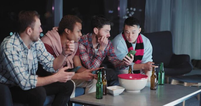 Diverse male company of cheerful friends watching football game and sharing smartphones browsing social media together staying home. Entertainment. Nomophobia.