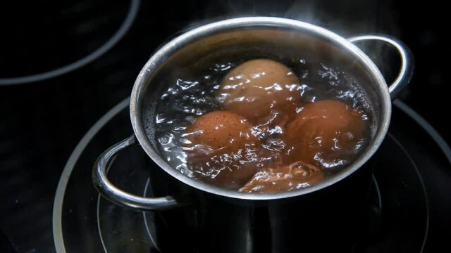 HD video. Close-up view of four brown chicken eggs boiling in hot water in small stainless saucepan on black kitchen stove (induction cooktop). Easter preparation theme.