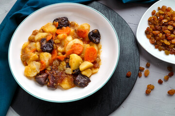 A dish of Jewish cuisine sweet tsimes with carrots vegetarian dates in a plate on a round stand on a concrete background next to raisins on a saucer.