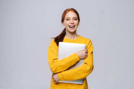 Smiling Young Woman Student Holding Laptop Computer And Looking At Camera On Isolated Gray Background. Pretty Lady Model With Red Hair Emotionally Showing Facial Expressions In Studio, Copy Space.