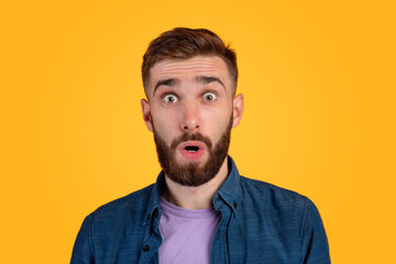 Unbelievable news. Portrait of bearded young guy opening mouth and eyes in shock, looking at camera on orange background