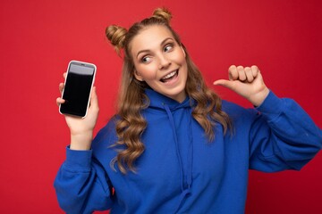 Attractive smiling young blonde woman wearing stylish blue hoodie isolated on red background with copy space holding smartphone showing phone in hand with empty screen display pointing at gadjet