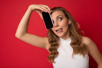 Naklejka premium Happy sexy attractive young blonde female person wearing white t-shirt isolated on red background with copy space holding smartphone showing phone in hand with empty screen for cutout looking to the