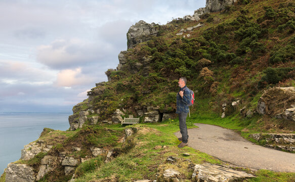 A hiker with a backpack stops on the coastal path and looks out to sea. The trail is in Somerset in the south of England. In the background are the Bristol Channel and a sky with clouds.