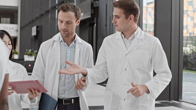 Medium shot of two male doctors going along hospital and talking. Then their Asian female colleague joining them and giving them digital tablet with important info
